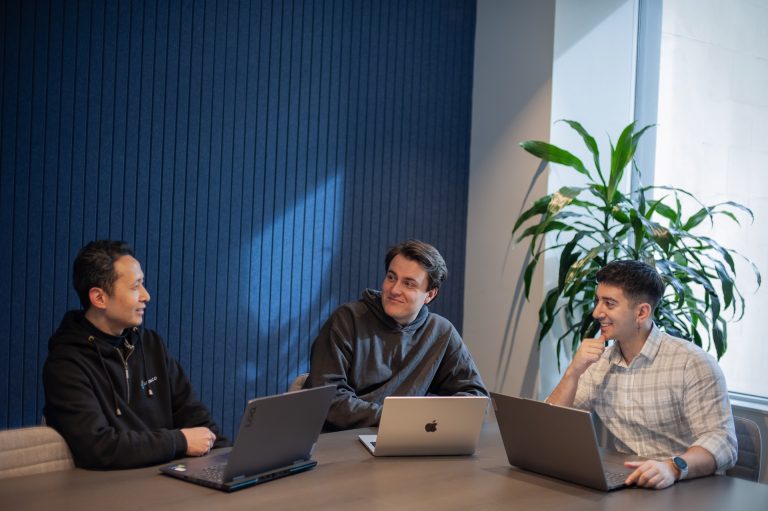3 people sat around a table with their laptops, talking.